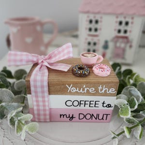 May include: A decorative wooden block set with the text "You're the COFFEE to my DONUT." The set is tied with a pink and white gingham ribbon. Miniature donuts and a coffee cup sit on top. A pink pitcher and house are in the background.