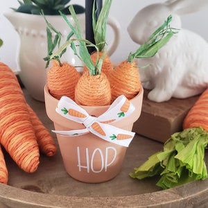 May include: A terracotta pot with a white ribbon and the word "HOP" written on it. The pot is filled with orange yarn carrots with green tops. The pot is sitting on a wooden tiered tray.