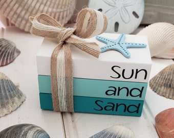 May include: A three-tiered wooden block set with a teal, light blue, and white paint finish. The blocks are stacked and tied together with a natural twine bow. The top block has a blue starfish and the text "Sun and Sand".