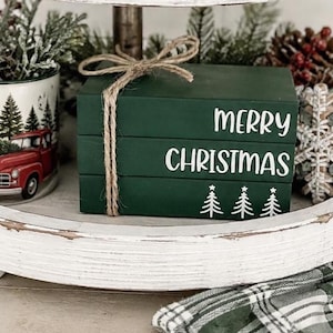 May include: A stack of three dark green wooden blocks tied with a natural jute twine bow. The top block features the white text MERRY, the middle block features the white text CHRISTMAS, and the bottom block features three white pine tree icons.