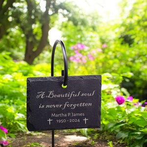 May include: A black slate garden marker with the inscription "A Beautiful soul is never forgotten" and the name Martha P. James, with dates 1950-2024. The marker is supported by a black metal arch and set in a garden with green foliage and pink flowers.