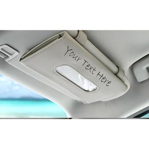 May include: A white leather car tissue holder with a clear plastic window for dispensing tissues. The holder is attached to the visor of a car and has the text "Your Text Here" written on it.