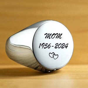 May include: A silver signet ring with a polished finish. The ring features the word "MOM" above the years "1956-2024" and two intertwined hearts. The ring is set against a warm, wooden background.