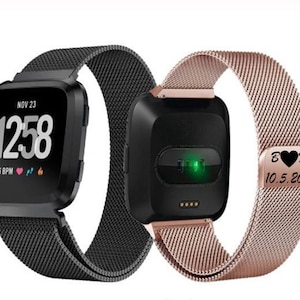 May include: Two smartwatches with black square faces and mesh bands. One watch displays the time, date, and heart rate. The other watch has a rose gold band and a small tag with initials and a date.