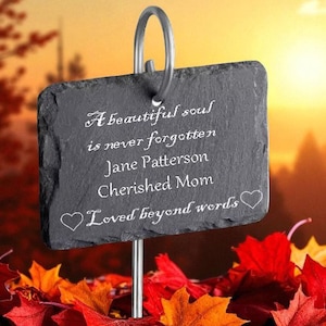 May include: A dark gray slate memorial plaque with white script, including the words "A beautiful soul is never forgotten" and "Loved beyond words." The plaque is mounted on a metal stake, surrounded by red and orange fall leaves.