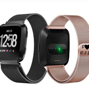 Puede incluir: Dos smartwatches con pantallas cuadradas negras y correas de malla. Uno muestra la fecha, hora y frecuencia card&iacute;aca. El otro tiene una correa de oro rosa con iniciales grabadas y una fecha. Accesorios para el seguimiento de la actividad f&iacute;sica.