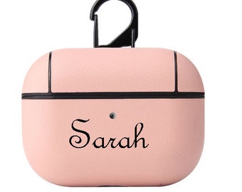 May include: A light pink AirPod case with the name "Sarah" in a cursive font. The case has a black border and a black carabiner clip. The case is designed to protect and carry wireless earbuds.