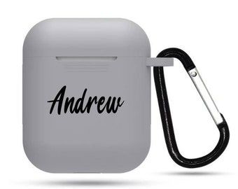 May include: A light gray AirPod case with the name "Andrew" in black script. The case has a black carabiner clip attached for easy carrying. The case is designed to protect and carry wireless earbuds.