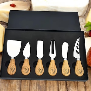 May include: Set of six cheese knives with wooden handles. Each handle has a personalized initial with a leaf design. The knives are in a black box with a foam insert.