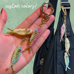 May include: Handmade beaded fish keychains in various colors, including pink, green, and cream. Each fish features intricate beadwork, gold-colored accents, and a keychain ring. The image includes the text "custom colors!"