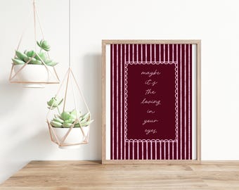May include: A framed print with the text "maybe it's the loving in your eyes" in a decorative font. The print has a burgundy background with a white scalloped border and is set against a white and burgundy striped background. Two hanging planters with succulents are also visible.