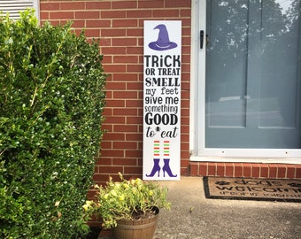May include: A white rectangular Halloween sign with the text "TRICK OR TREAT SMELL my feet give me something GOOD to eat". The sign features a purple witch hat and boots. The sign is placed next to a green bush and a flower pot.