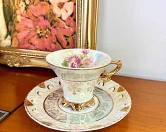 May include: A vintage teacup and saucer set. The teacup features a floral design with pink and purple flowers, gold trim, and a gold handle. The saucer has a matching design with gold accents. The set is displayed on a wooden surface.