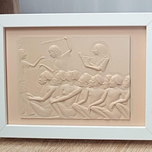 Könnte beinhalten: Gerahmte Reliefskulptur, die eine altägyptische Szene darstellt. Das Kunstwerk in Beige- und Brauntönen zeigt Figuren in verschiedenen Posen, einige sitzen und andere stehen, mit detaillierten Schnitzereien und einem weißen Rahmen.
