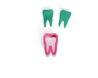 May include: A pink plastic tooth-shaped cookie cutter with two green plastic teeth on a white background.