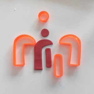 May include: A set of five cookie cutters in orange and red. The cutters are shaped like a circle, a rectangle, and two curved shapes. The cutters are on a white surface.
