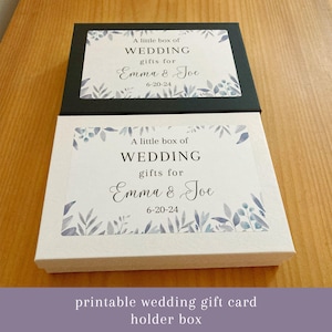 May include: Two rectangular gift boxes, one black and one white, are stacked on a wooden surface. Each box has a printed label with the text "A little box of WEDDING gifts for Emma & Joe 6-20-24" and floral accents. The boxes are for wedding gifts.