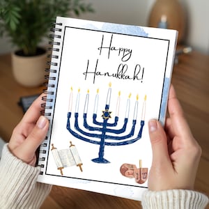 May include: A spiral-bound notebook featuring a white cover with the text Happy Hanukkah! in black script. The design includes a blue menorah with yellow flames, a Star of David, a scroll, and a dreidel, all set against a light blue watercolor wash.