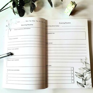 Puede incluir: Diario abierto con indicaciones para rutinas matutinas y vespertinas. Las p&aacute;ginas blancas presentan texto negro y dise&ntilde;os florales. Incluye indicaciones para afirmaciones, intenciones, gratitud y autocuidado. Tambi&eacute;n se ve un bol&iacute;grafo y cinta decorativa.