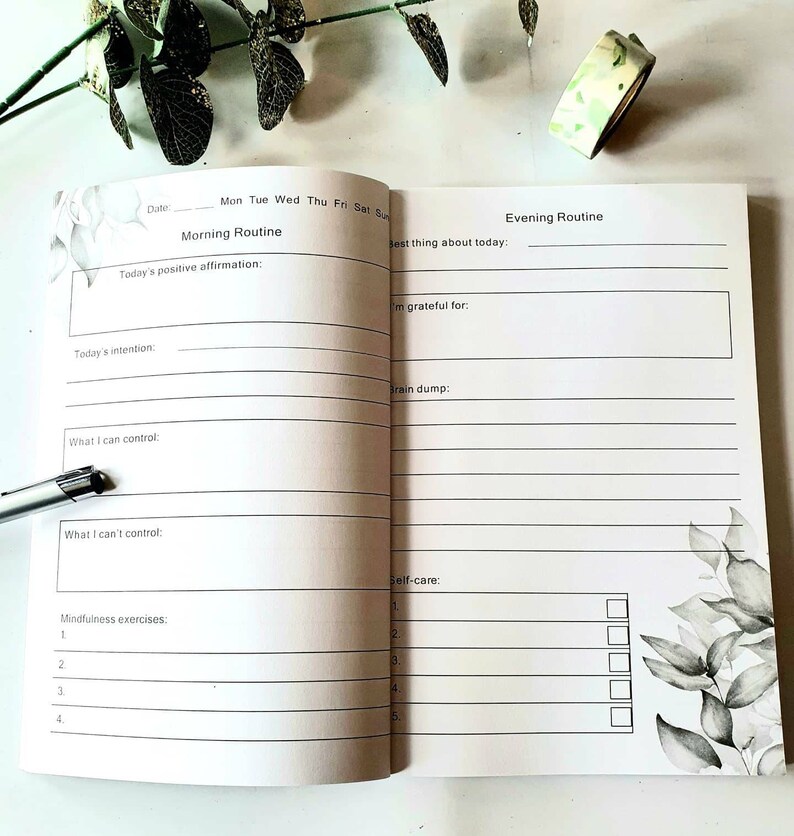 Puede incluir: Diario abierto con indicaciones para rutinas matutinas y vespertinas. Las p&aacute;ginas blancas presentan texto negro y dise&ntilde;os florales. Incluye indicaciones para afirmaciones, intenciones, gratitud y autocuidado. Tambi&eacute;n se ve un bol&iacute;grafo y cinta decorativa.