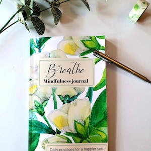 Puede incluir: Un diario de mindfulness con un dise&ntilde;o floral con flores blancas y hojas verdes. La portada dice "Breathe Mindfulness journal" y "Daily practices for a happier you". Tambi&eacute;n hay un bol&iacute;grafo plateado y cinta adhesiva decorativa.