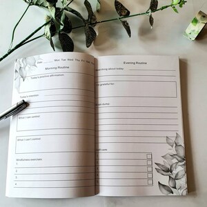 Puede incluir: Diario abierto con indicaciones de rutina matutina y vespertina. Las p&aacute;ginas del diario son blancas con detalles florales grises. Un bol&iacute;grafo plateado descansa sobre la p&aacute;gina. El texto incluye indicaciones para afirmaciones, intenciones, gratitud y autocuidado.