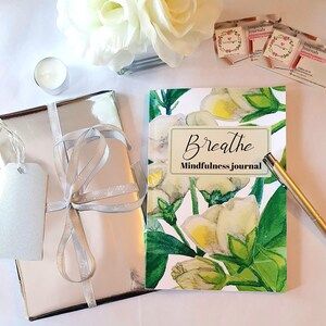 Puede incluir: Un regalo envuelto en plata con una cinta plateada y una etiqueta. Un diario con un dise&ntilde;o floral y la palabra "Breathe" est&aacute; junto a un bol&iacute;grafo dorado. Tambi&eacute;n hay una peque&ntilde;a vela y una flor blanca.