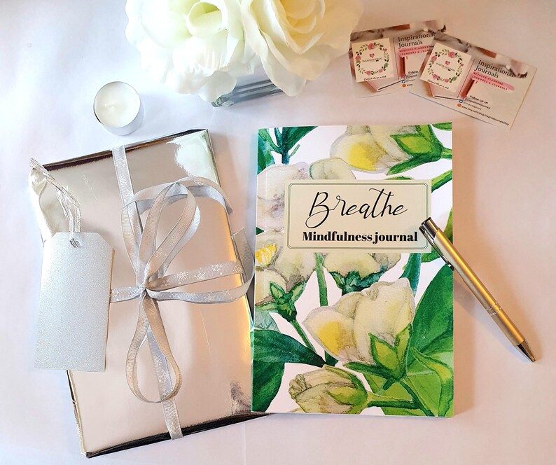 Puede incluir: Un regalo envuelto en plata con una cinta plateada y una etiqueta. Un diario con un dise&ntilde;o floral y la palabra "Breathe" est&aacute; junto a un bol&iacute;grafo dorado. Tambi&eacute;n hay una peque&ntilde;a vela y una flor blanca.