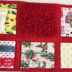 Puede incluir: Una manta sensorial roja y blanca con varias texturas y patrones. La manta presenta una cremallera, un cuadrado rojo peludo, una borla blanca, una tela blanca con corazones rojos, una tela blanca con dinosaurios, una tela blanca con un diseño de princesa y una tela blanca con un patrón de puntos blancos.