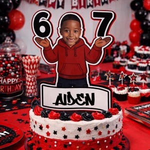 Puede incluir: Una tarta de cumpleaños con glaseado rojo, negro y blanco, coronada con un recorte de dibujos animados de un niño sosteniendo los números 6 y 7. La tarta tiene un letrero que dice "Aiden". Globos y cupcakes en el fondo.