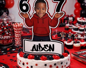 May include: A birthday cake with red, black, and white frosting, topped with a cartoon cutout of a boy holding the numbers 6 and 7. The cake has a sign that says "Aiden". The background includes balloons and cupcakes.