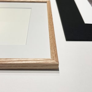 May include: A light-colored wooden picture frame with a white mat board border. The frame has a visible wood grain texture and a rectangular shape. A second black picture frame with a white mat board is partially visible in the background.