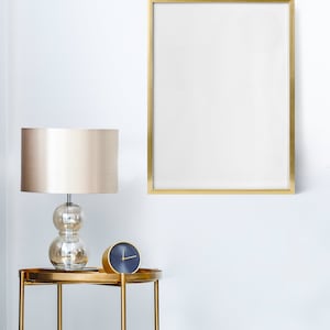 May include: A gold-framed picture sits above a gold side table with a cream lampshade and a blue clock.