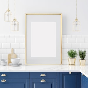 May include: A gold framed picture with a white mat and a white background hangs on a white tiled wall above a blue kitchen cabinet with gold handles. There are three gold pendant lights hanging above the picture.