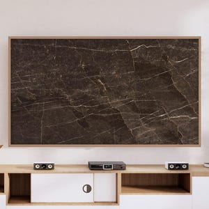 May include: A large, rectangular dark brown marble panel with white veining is mounted on a wall. The panel is framed in light brown wood. Below, a light wood and white cabinet holds electronics and decorative items.