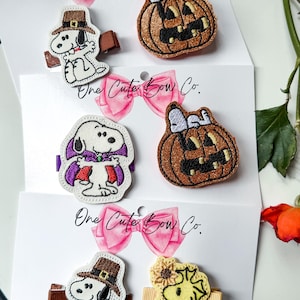 May include: Six hair clips featuring embroidered Snoopy and Woodstock characters on white display cards labeled One Cute Bow Co. Designs include Snoopy as a pilgrim, a vampire, and sitting on a glittery orange pumpkin, plus Woodstock with a sunflower.