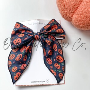 May include: A large hair bow with a Halloween theme. The bow is navy blue with a pattern of orange jack-o'-lanterns and stars. The bow is attached to a white card with the text "one cute bow co" and "onecutebowcoetsy.com".