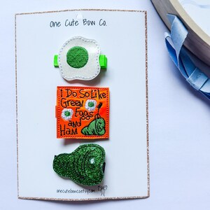 May include: A set of three hair clips with a Dr. Seuss theme. The clips include a fried egg, a square with the text "I Do So Like Green Eggs and Ham", and a green ham. The clips are attached to a white card with the text "one Cute Bow Co."