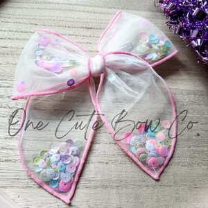 Op de afbeelding: Een witte tule strik met roze bies en kleurrijke, iriserende pailletten. De strik is in een knoop geknoopt en heeft een lus aan de bovenkant.