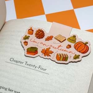 Könnte beinhalten: Ein rosa Lesezeichen mit herbstlichen Illustrationen, darunter ein Kürbis, Blätter und eine Kerze. Der Text "I'm an autumn" steht auf dem Lesezeichen. Das Lesezeichen befindet sich in einem offenen Buch.