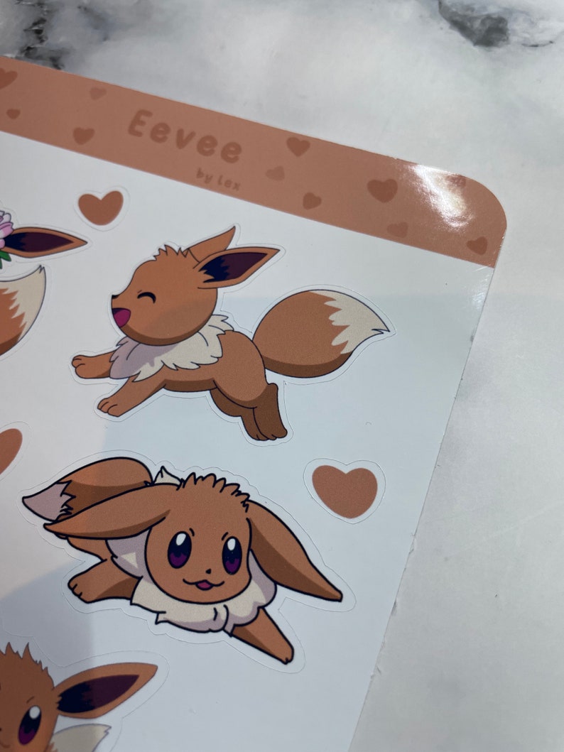 K&ouml;nnte beinhalten: Ein Blatt mit braunen und wei&szlig;en Aufklebern mit einem Cartoon-Eevee, einem Pok&eacute;mon-Charakter. Die Aufkleber sind mit Herzen und dem Text "Eevee by lex" verziert.