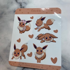 K&ouml;nnte beinhalten: Ein Blatt mit braunen und wei&szlig;en Aufklebern mit f&uuml;nf verschiedenen Illustrationen von Eevee, einem Pok&eacute;mon-Charakter. Die Aufkleber sind von kleinen braunen Herzen umgeben. Der Text "Eevee by lex" und "made with love" ist auf dem Blatt gedruckt.