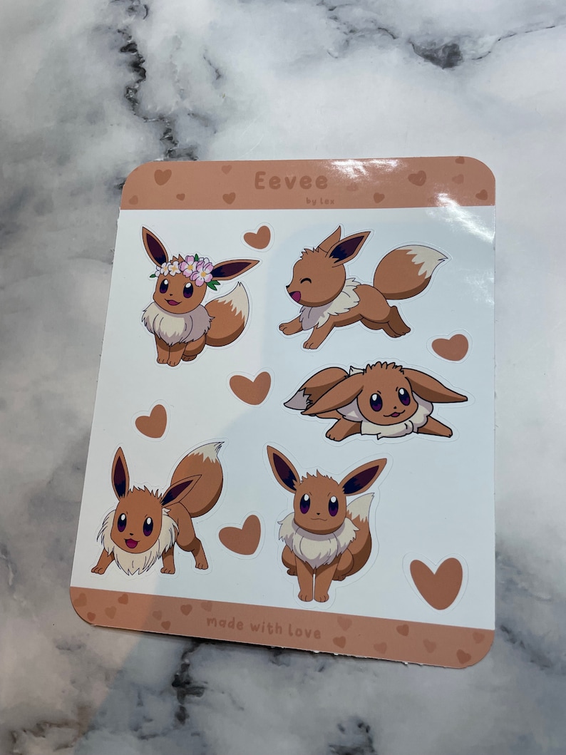 K&ouml;nnte beinhalten: Ein Blatt mit braunen und wei&szlig;en Aufklebern mit f&uuml;nf verschiedenen Illustrationen von Eevee, einem Pok&eacute;mon-Charakter. Die Aufkleber sind von kleinen braunen Herzen umgeben. Der Text "Eevee by lex" und "made with love" ist auf dem Blatt gedruckt.