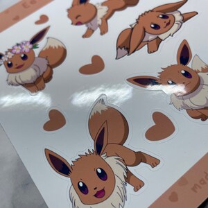 K&ouml;nnte beinhalten: Ein Bogen mit Aufklebern, die verschiedene Posen von Eevee zeigen, einem braunen fuchsartigen Pok&eacute;mon mit einem buschigen Schwanz. Die Aufkleber befinden sich auf einem wei&szlig;en Hintergrund mit braunen Herzen.