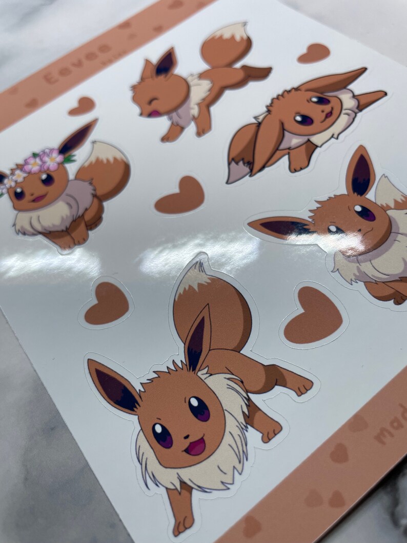 K&ouml;nnte beinhalten: Ein Bogen mit Aufklebern, die verschiedene Posen von Eevee zeigen, einem braunen fuchsartigen Pok&eacute;mon mit einem buschigen Schwanz. Die Aufkleber befinden sich auf einem wei&szlig;en Hintergrund mit braunen Herzen.