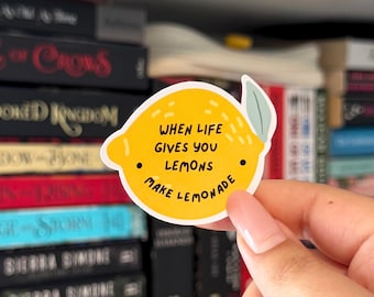 May include: A yellow lemon-shaped sticker with the text "WHEN LIFE GIVES YOU LEMONS MAKE LEMONADE" in black. The sticker has a green leaf and two black dots. The sticker is held in front of a bookshelf filled with books.