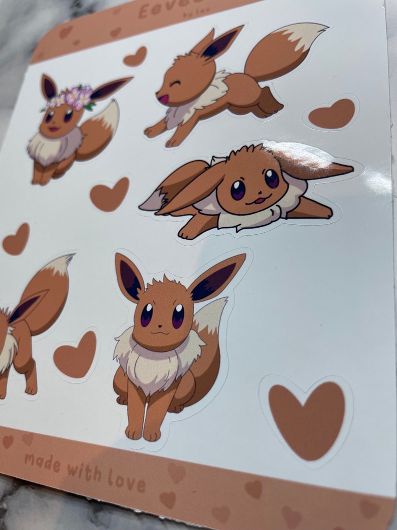 K&ouml;nnte beinhalten: Ein Blatt mit braunen und wei&szlig;en Aufklebern mit verschiedenen Illustrationen von Eevee, einem Pok&eacute;mon-Charakter. Die Aufkleber befinden sich auf einem wei&szlig;en Hintergrund mit braunem Rand und dem Text "made with love".