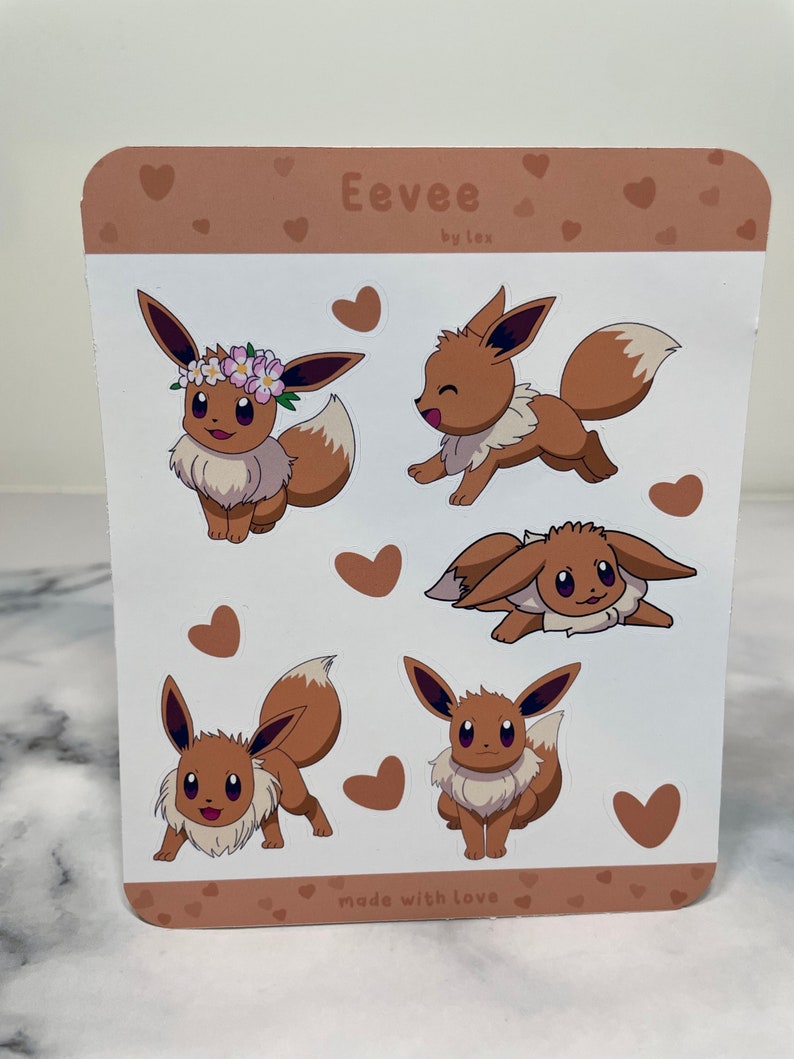 K&ouml;nnte beinhalten: F&uuml;nf braune und wei&szlig;e Eevee Pokemon-Aufkleber auf einem hellbraunen Hintergrund mit Herzen. Die Aufkleber zeigen Eevee in verschiedenen Posen, darunter eine mit einer Blumenkrone. Der Text "Eevee by Lex" und "made w/th love" befindet sich oben und unten auf dem Blatt.