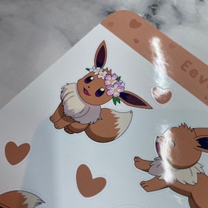 K&ouml;nnte beinhalten: Ein Set mit braunen und wei&szlig;en Eevee-Aufklebern mit einer Blumenkrone. Die Aufkleber befinden sich auf einem wei&szlig;en Hintergrund mit braunen Herzen.