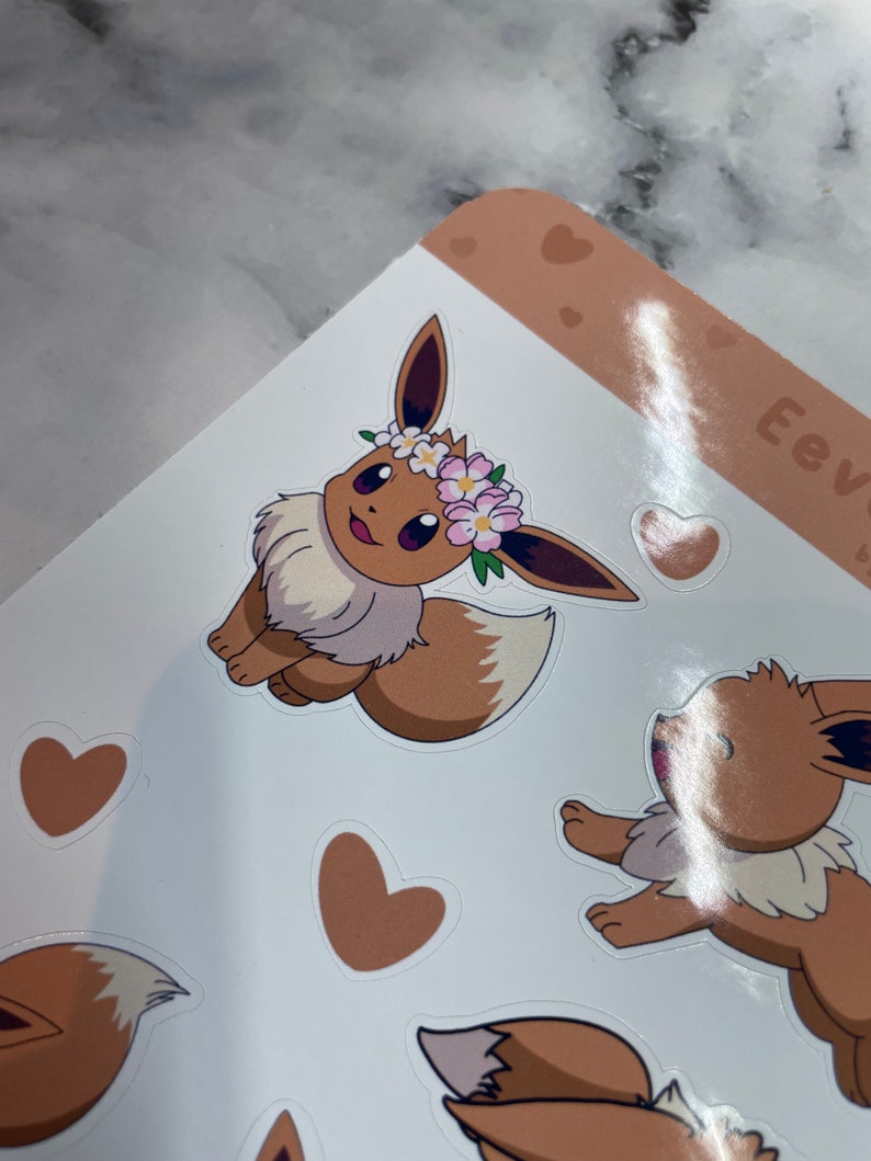 K&ouml;nnte beinhalten: Ein Set mit braunen und wei&szlig;en Eevee-Aufklebern mit einer Blumenkrone. Die Aufkleber befinden sich auf einem wei&szlig;en Hintergrund mit braunen Herzen.